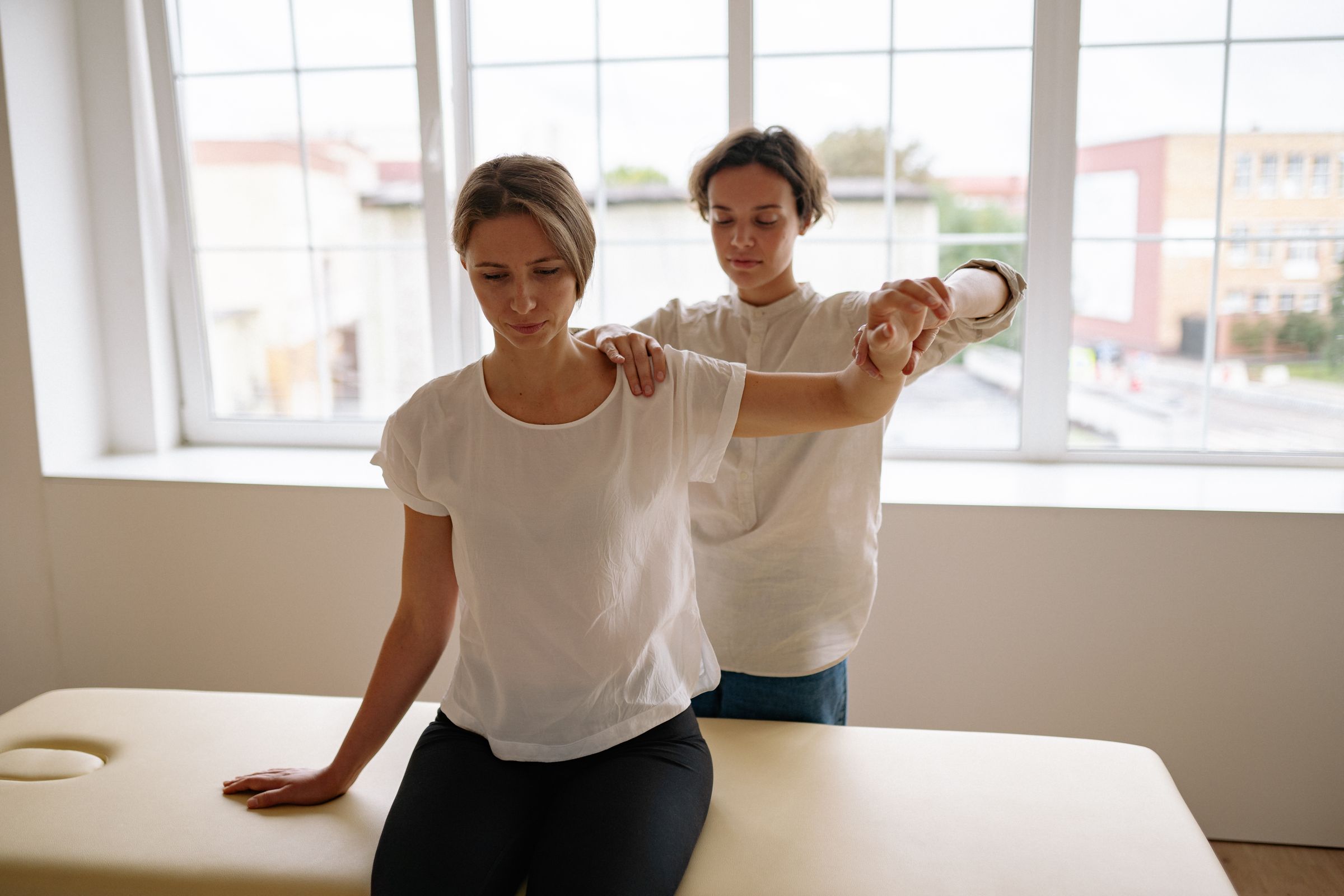 Physiotherapist guiding a seated patient through an assisted shoulder stretch in a bright, modern clinic